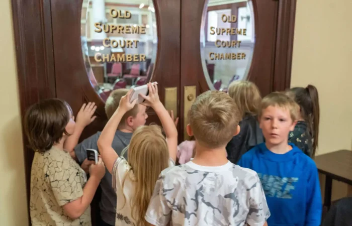 Secretary of State's Office, Governor's Office meet with Warren Elementary 2nd graders 9 A group of children stand in front of wooden double doors labeled Old Supreme Court Chamber, with some peering through the glass and others holding devices, possibly taking pictures or videos.