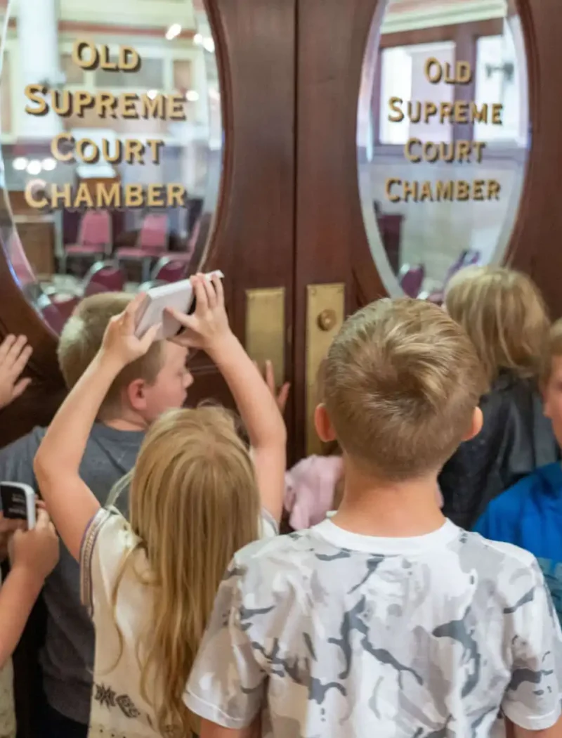 A group of children stand in front of wooden double doors labeled Old Supreme Court Chamber, with some peering through the glass and others holding devices, possibly taking pictures or videos.