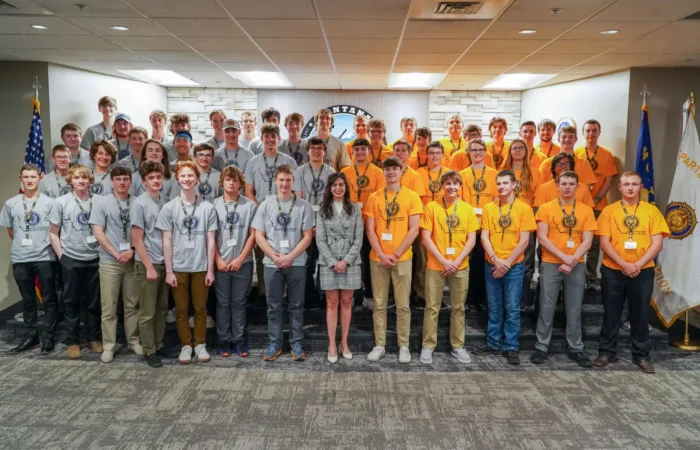 Secretary of State Christi Jacobsen a featured speaker at American Legion Boys State 2022 9 A large group of young men in gray and yellow T-shirts pose with Secretary of State Christi Jacobsen indoors, standing in rows in front of flags and a sign that says Montana at American Legion Boys State.