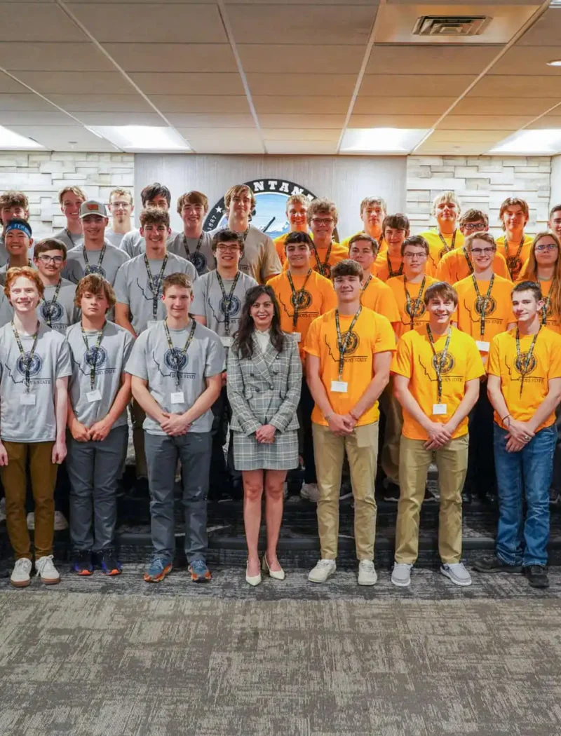 Secretary of State Christi Jacobsen a featured speaker at American Legion Boys State 2022 2 A large group of young men in gray and yellow T-shirts pose with Secretary of State Christi Jacobsen indoors, standing in rows in front of flags and a sign that says Montana at American Legion Boys State.