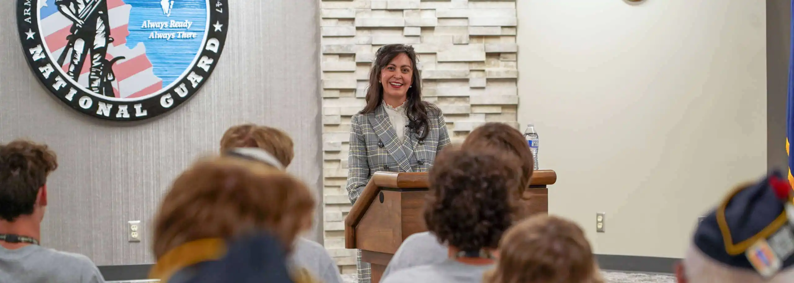 Secretary of State Christi Jacobsen stands at a podium, smiling as she addresses the 2022 American Legion Boys State attendees. Behind her, the National Guard emblem is displayed while the audience listens attentively.