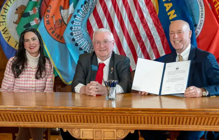 Secretary Christi Jacobsen, Governor Gianforte sign bill delivering photo ID for voting in Montana 4 Three people sit at a wooden table in front of U.S. and Montana flags; Secretary Christi Jacobsen, on the left, Senator Mike Cuffe in the center, and Governor Greg Gianforte on the right holds up a signed photo ID for voting document as all three smile at the camera.