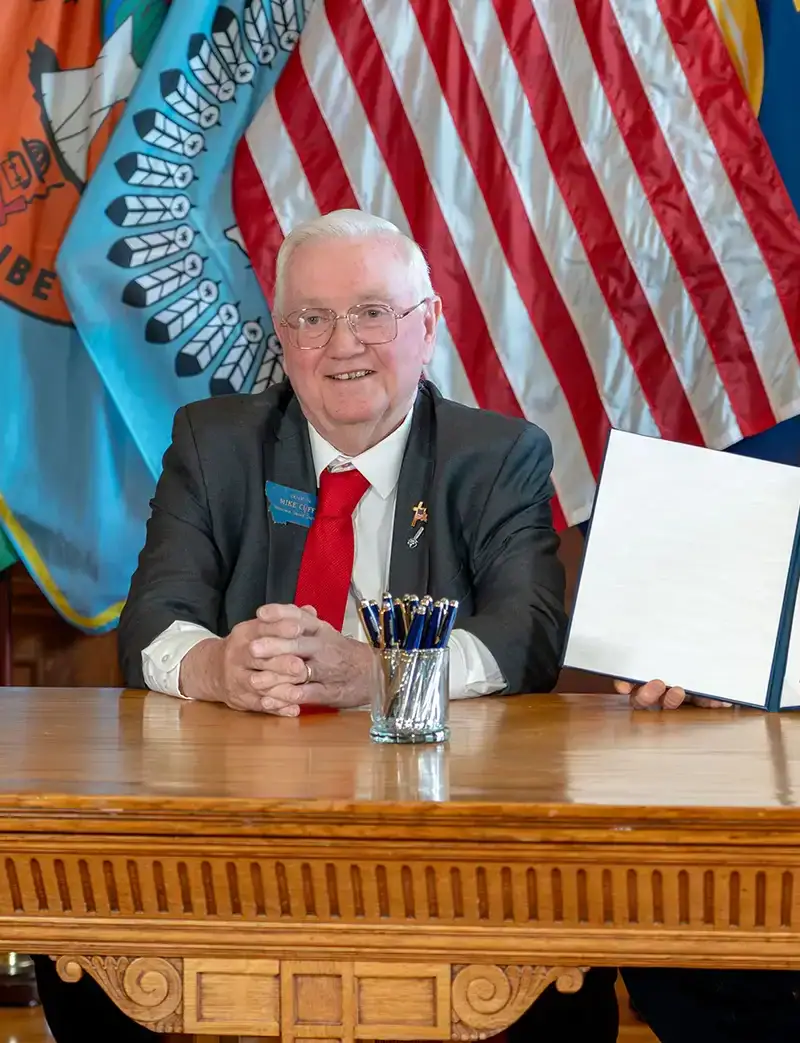 Three people sit at a wooden table in front of U.S. and Montana flags; Secretary Christi Jacobsen, on the left, Senator Mike Cuffe in the center, and Governor Greg Gianforte on the right holds up a signed photo ID for voting document as all three smile at the camera.