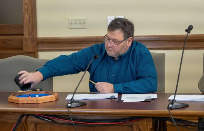 Secretary Christi Jacobsen: 'Board of State Canvassers roll the dice for 2024 Primary post-election audit selection' 8 A man seated at a table draws an item from a black container during a post-election audit, with papers and a wooden tray in front of him. He is wearing glasses and a teal shirt, and a microphone is on the table.