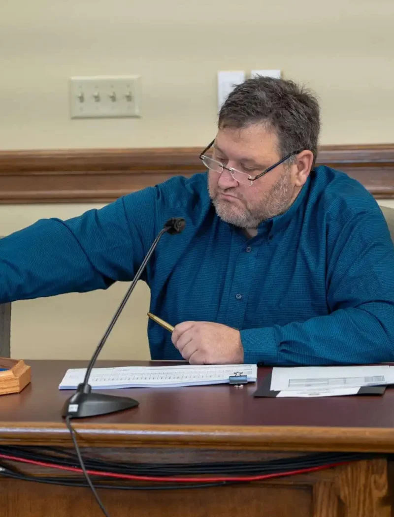 A man seated at a table draws an item from a black container during a post-election audit, with papers and a wooden tray in front of him. He is wearing glasses and a teal shirt, and a microphone is on the table.