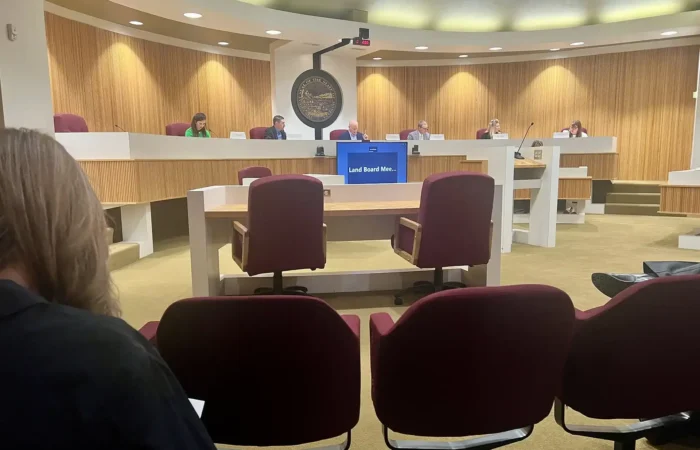 A meeting is taking place in a government or council chamber with people seated at a curved desk facing the audience. The room, with wood-paneled walls, red chairs, and a large seal, is set to discuss logging and oil and gas projects.