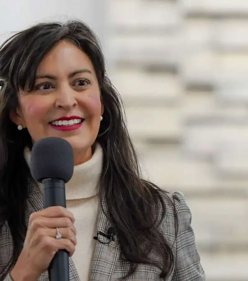 A woman with long dark hair, wearing a plaid blazer and turtleneck, smiles while speaking into a microphone. In the background, a blurred stone wall and part of a round sign hint at Montana businesses and 2025 Annual Report fees discussion.