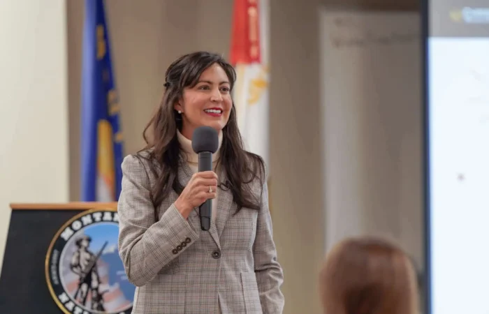 Secretary Christi Jacobsen helps commence 2024 Montana American Legion Boys & Girls State 2 A woman in a plaid blazer holds a microphone and speaks at a Montana American Legion Girls State event, standing near a podium with the Montana state seal and flags. A presentation screen is partially visible in the background.
