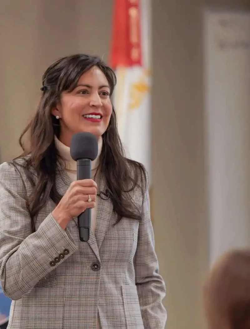 A woman in a plaid blazer holds a microphone and speaks at a Montana American Legion Girls State event, standing near a podium with the Montana state seal and flags. A presentation screen is partially visible in the background.