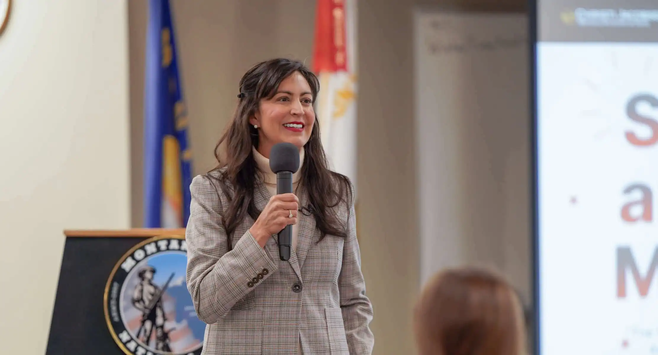 A woman in a plaid blazer holds a microphone and speaks at a Montana American Legion Girls State event, standing near a podium with the Montana state seal and flags. A presentation screen is partially visible in the background.