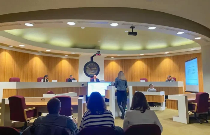 People sit and observe as a woman stands and speaks at a podium in a modern courtroom or council chamber, with officials—including Land Board member Christi Jacobsen—seated at a raised desk, and a large screen displaying information on Montana schools.