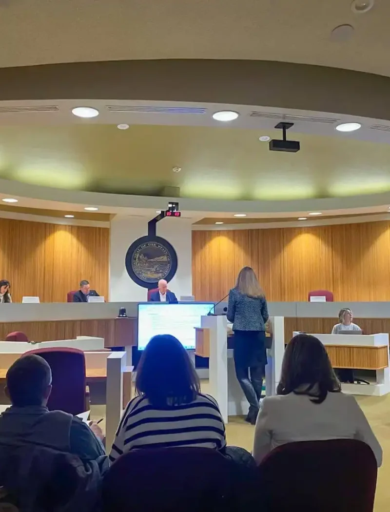 People sit and observe as a woman stands and speaks at a podium in a modern courtroom or council chamber, with officials—including Land Board member Christi Jacobsen—seated at a raised desk, and a large screen displaying information on Montana schools.