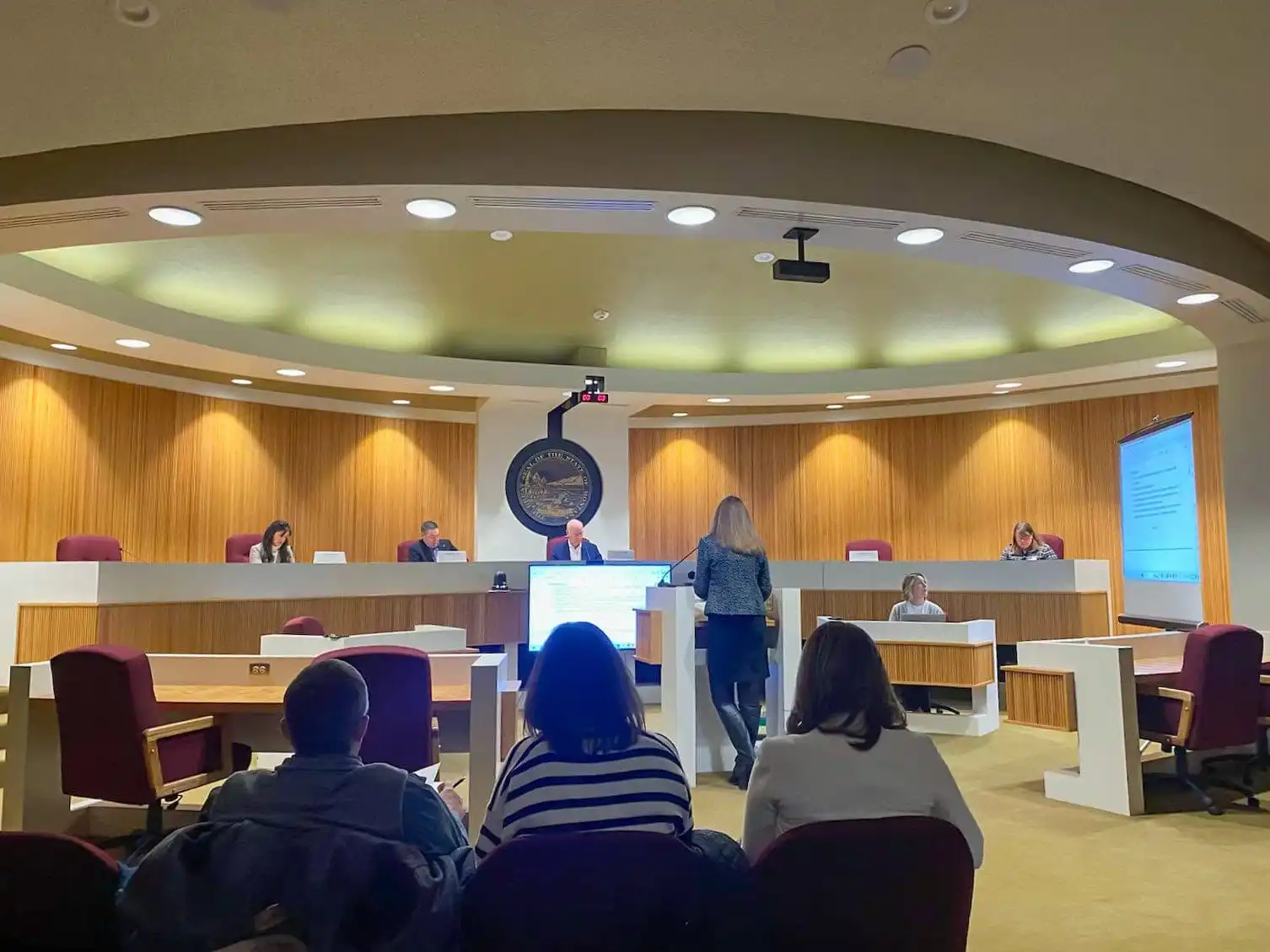 People sit and observe as a woman stands and speaks at a podium in a modern courtroom or council chamber, with officials—including Land Board member Christi Jacobsen—seated at a raised desk, and a large screen displaying information on Montana schools.