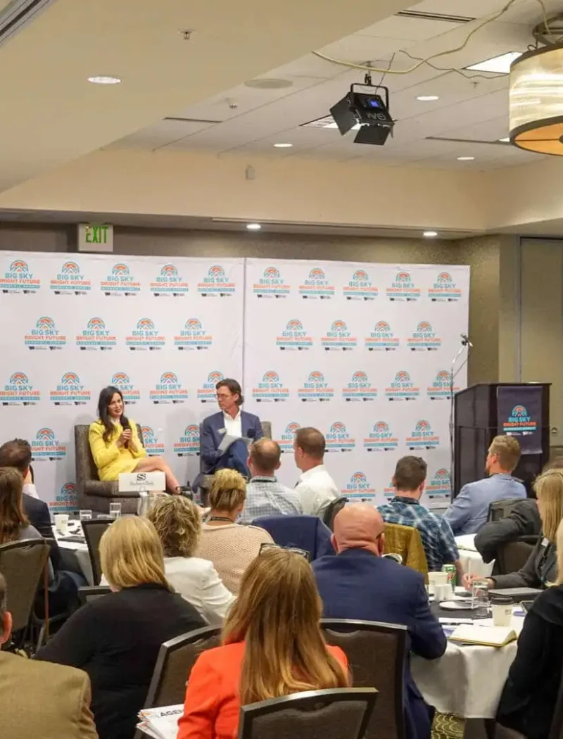 A conference room filled with attendees seated at round tables, listening to two speakers on stage in front of a branded Chamber of Commerce backdrop. People are focused on the presentation, with screens and lights visible in the room.