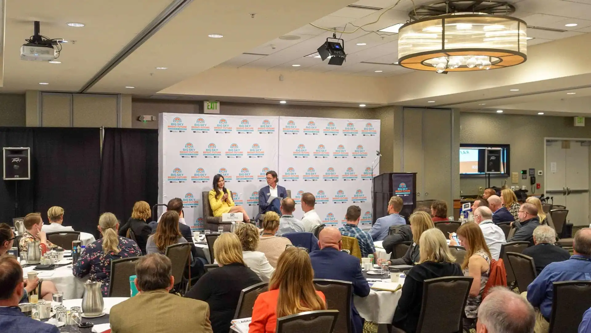 A conference room filled with attendees seated at round tables, listening to two speakers on stage in front of a branded Chamber of Commerce backdrop. People are focused on the presentation, with screens and lights visible in the room.