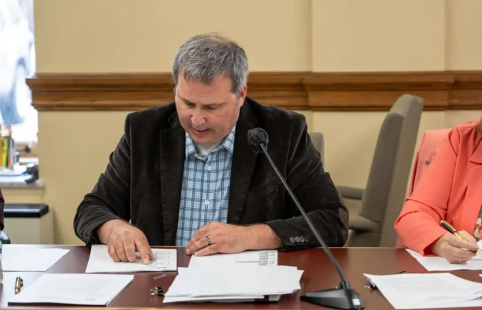 Secretary Christi Jacobsen: 'State Canvass completed for Montana's 2024 Primary Election' 7 A man in a dark blazer and plaid shirt sits at a table with papers, looking down and pointing at a document. A microphone and more papers are in front of him, and another person sits partially visible at his side.
