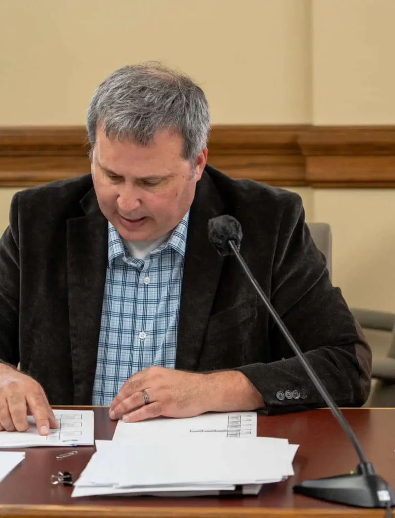 A man in a dark blazer and plaid shirt sits at a table with papers, looking down and pointing at a document. A microphone and more papers are in front of him, and another person sits partially visible at his side.