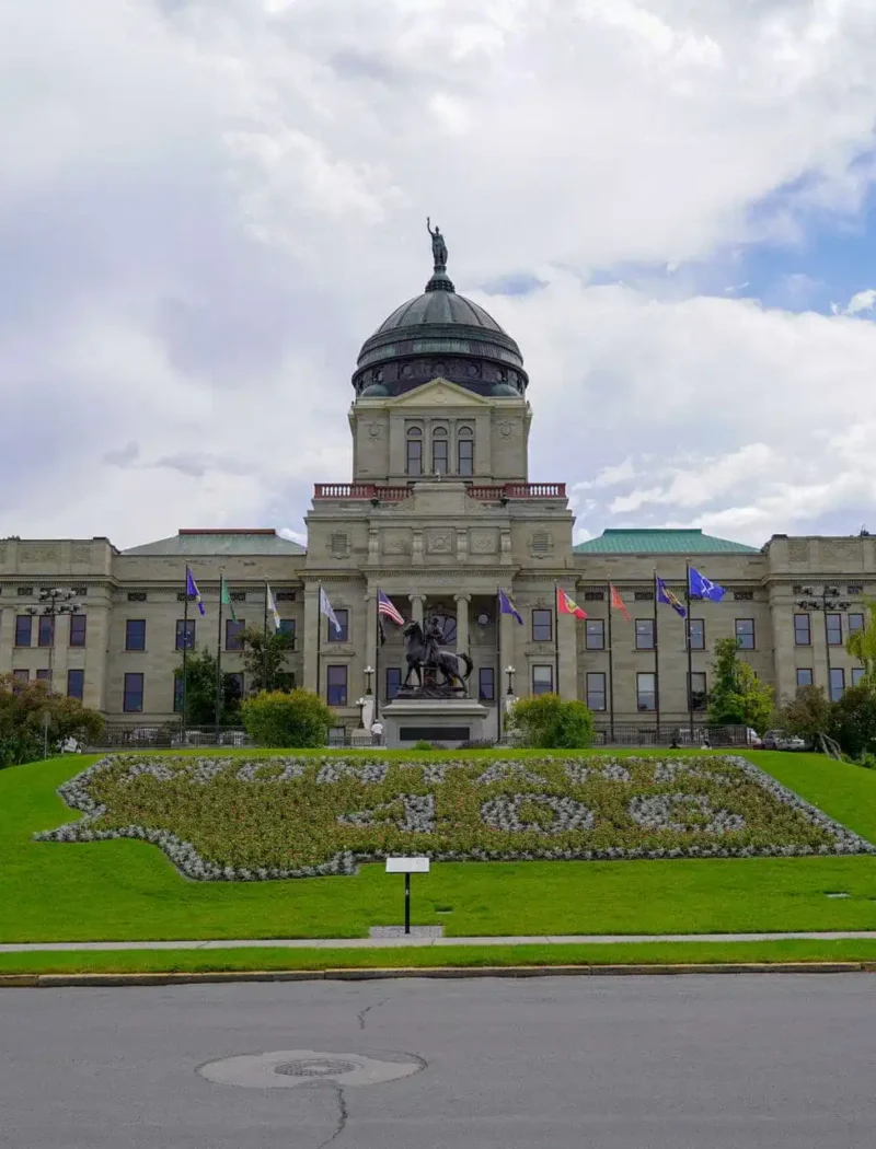 The Montana State Capitol building with its domed roof stands proudly amid green lawns and state flags, while a floral design graces the grass in front—capturing the spirit reflected during the 2022 Primary Election.