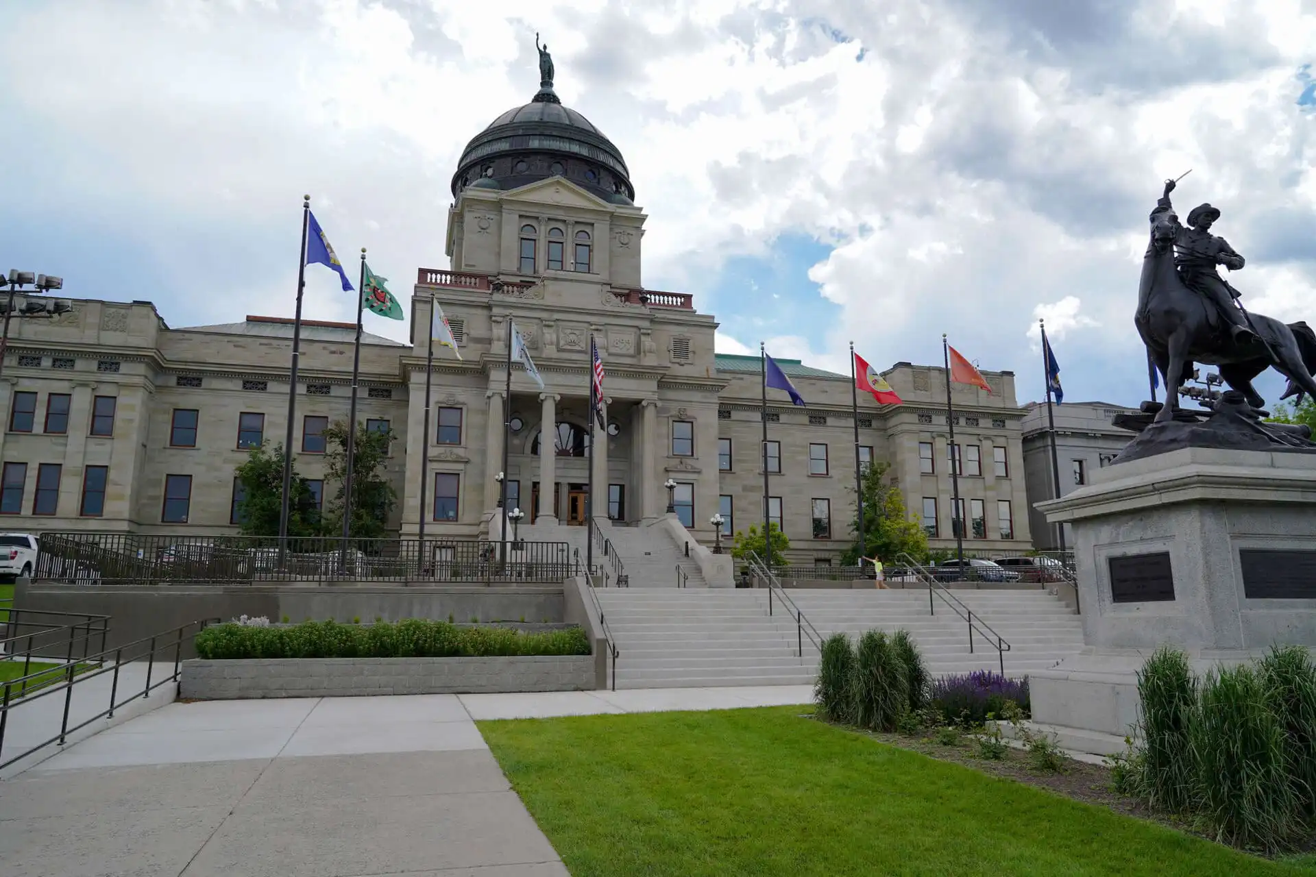 The image shows the Montana State Capitol building with a domed roof, multiple flags out front, and a horse and rider statue in the foreground—a landmark often visited by local election officials during tabletop exercise events.