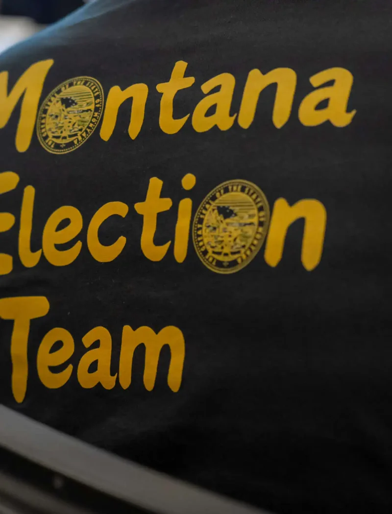 A person wearing a black shirt with yellow text that reads Montana Election Team and features two official state seals, supporting Secretary of State Christi Jacobsen on Primary Election Day.