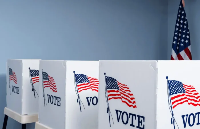 Secretary Jacobsen announces opening of polls for Primary Election 9 Three voting booths with the word VOTE and an American flag printed on each, set up in a row in a room with light blue walls; a U.S. flag stands in the background, marking the Primary Election and opening of polls announced by Secretary Jacobsen.