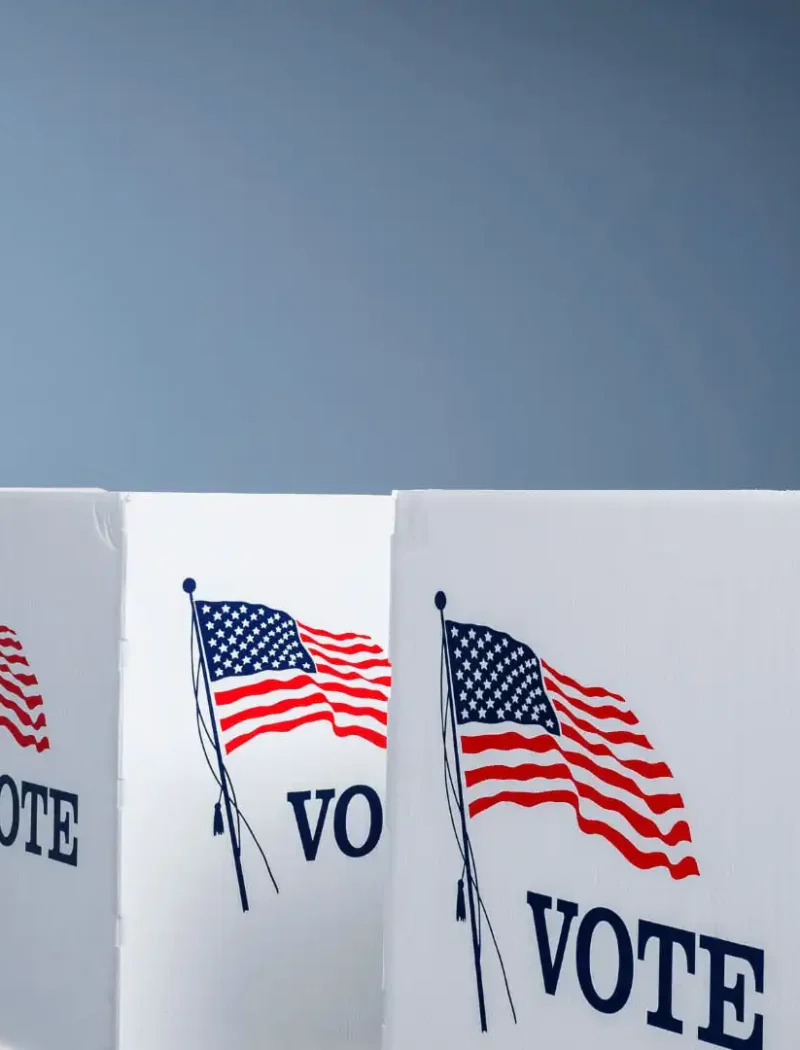 Secretary Jacobsen announces opening of polls for Primary Election 1 Three voting booths with the word VOTE and an American flag printed on each, set up in a row in a room with light blue walls; a U.S. flag stands in the background, marking the Primary Election and opening of polls announced by Secretary Jacobsen.