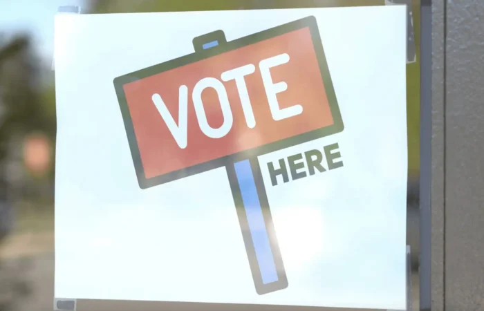 Secretary Jacobsen congratulates candidates, thanks Montana voters and election workers 8 A sign with a red and blue post reads VOTE HERE in bold letters, displayed on a window or door, guiding Montana voters to their polling location, where election workers assist with the voting process.