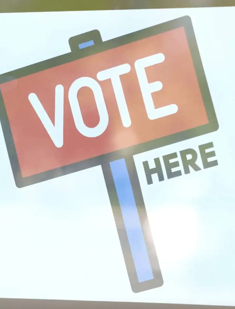 A sign with a red and blue post reads VOTE HERE in bold letters, displayed on a window or door, guiding Montana voters to their polling location, where election workers assist with the voting process.