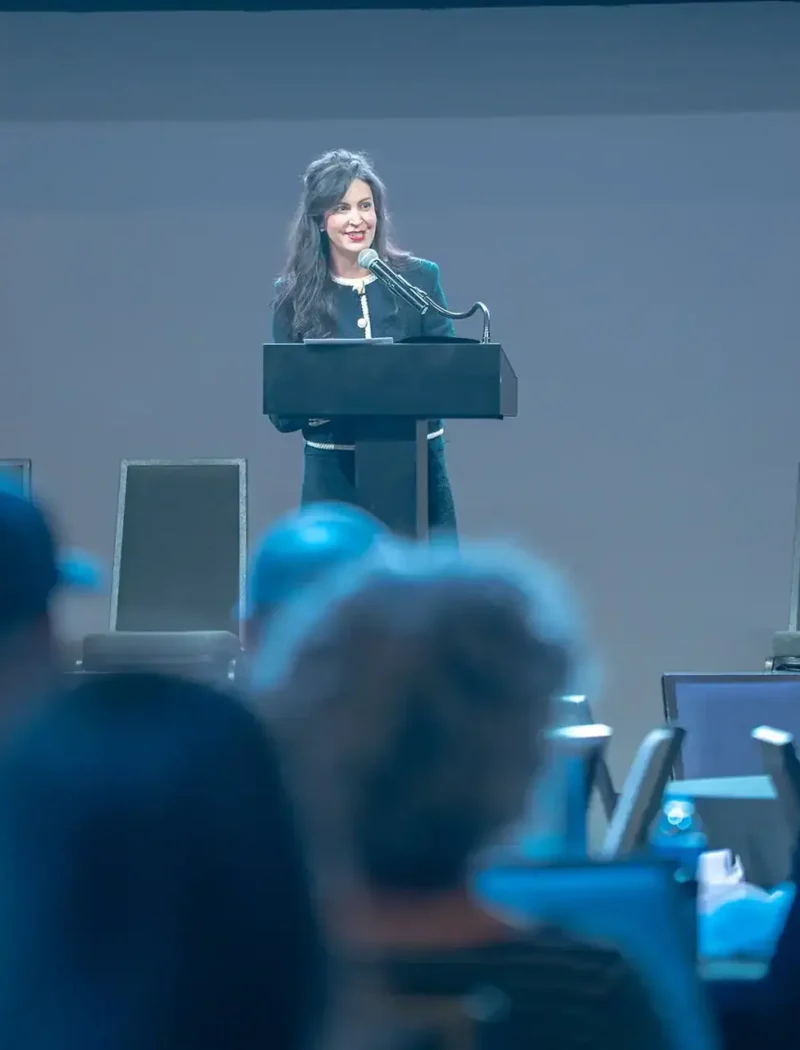 Christi Jacobsen stands and speaks at a podium on stage, smiling at the audience during a Montana Mining Association event. Several empty chairs are visible in the conference setting, highlighting her role in the Montana mining industry.