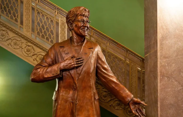 Secretary Christi Jacobsen joins Montanans in celebrating statue unveiling for Governor Judy Martz 10 A bronze statue of a smiling person in glasses, wearing a suit, with one hand on their chest and the other arm outstretched stands before an ornate staircase and a green wall, celebrating the statue unveiling of Governor Judy Martz.