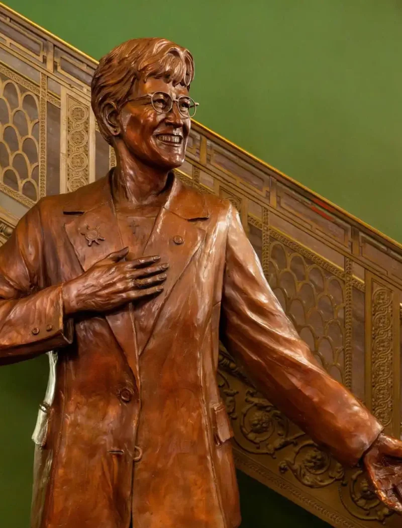 A bronze statue of a smiling person in glasses, wearing a suit, with one hand on their chest and the other arm outstretched stands before an ornate staircase and a green wall, celebrating the statue unveiling of Governor Judy Martz.