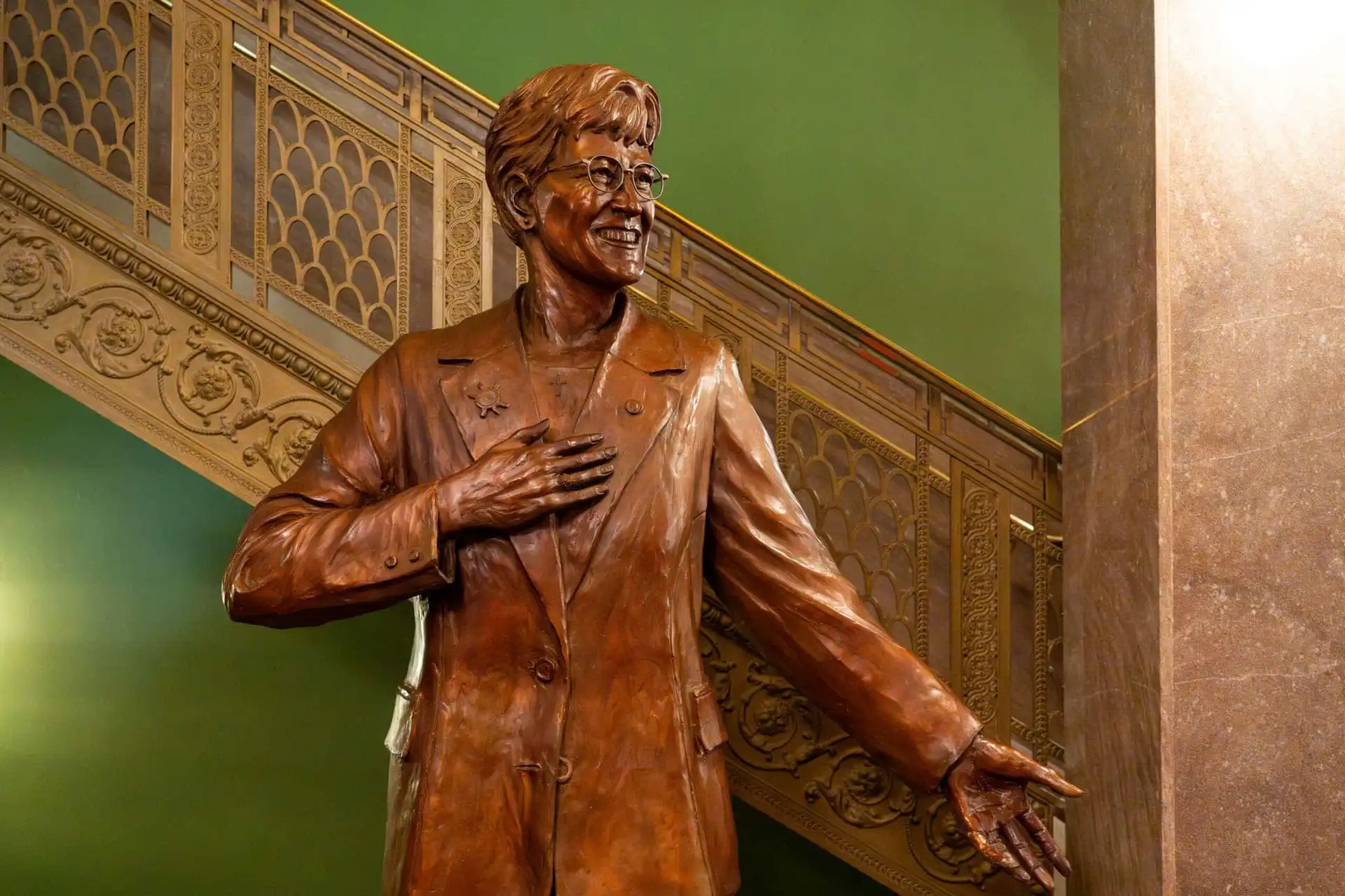A bronze statue of a smiling person in glasses, wearing a suit, with one hand on their chest and the other arm outstretched stands before an ornate staircase and a green wall, celebrating the statue unveiling of Governor Judy Martz.