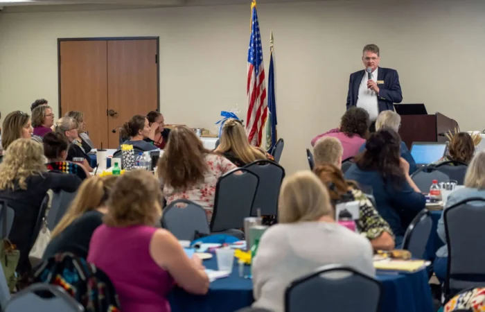 Montana Secretary of State's Office presents at 2022 Montana Association of Clerks & Recorders Convention 5 A man in a suit speaks at a podium to attendees of the 2022 Convention for the Montana Association of Clerks & Recorders. U.S. flags stand behind him as the audience listens attentively, some taking notes.