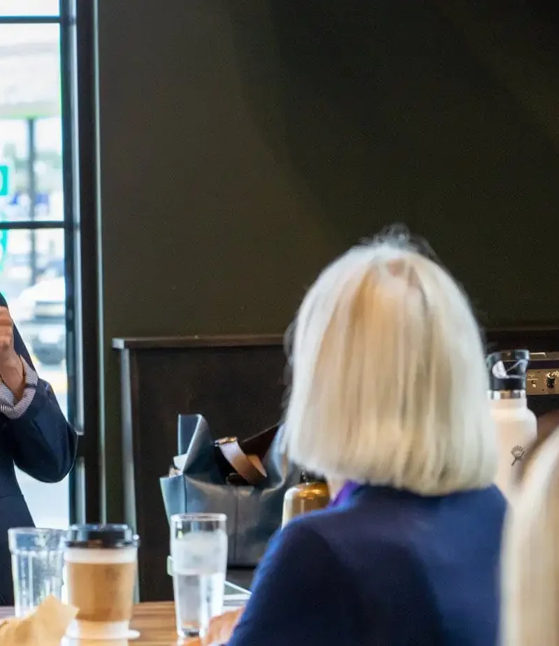 A woman in a suit speaks into a microphone and smiles while addressing a small group seated at a table in a brightly lit café or restaurant.