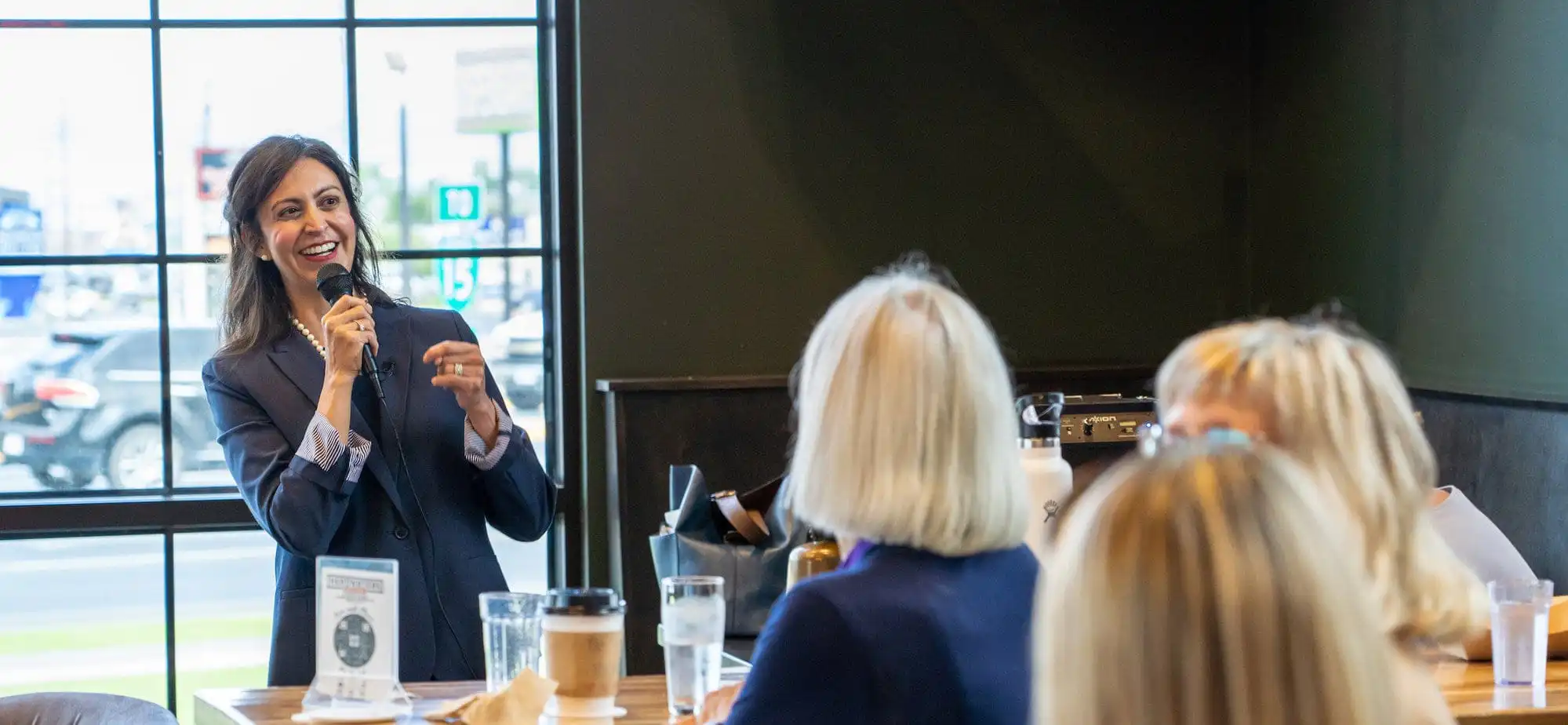 A woman in a suit speaks into a microphone and smiles while addressing a small group seated at a table in a brightly lit café or restaurant.
