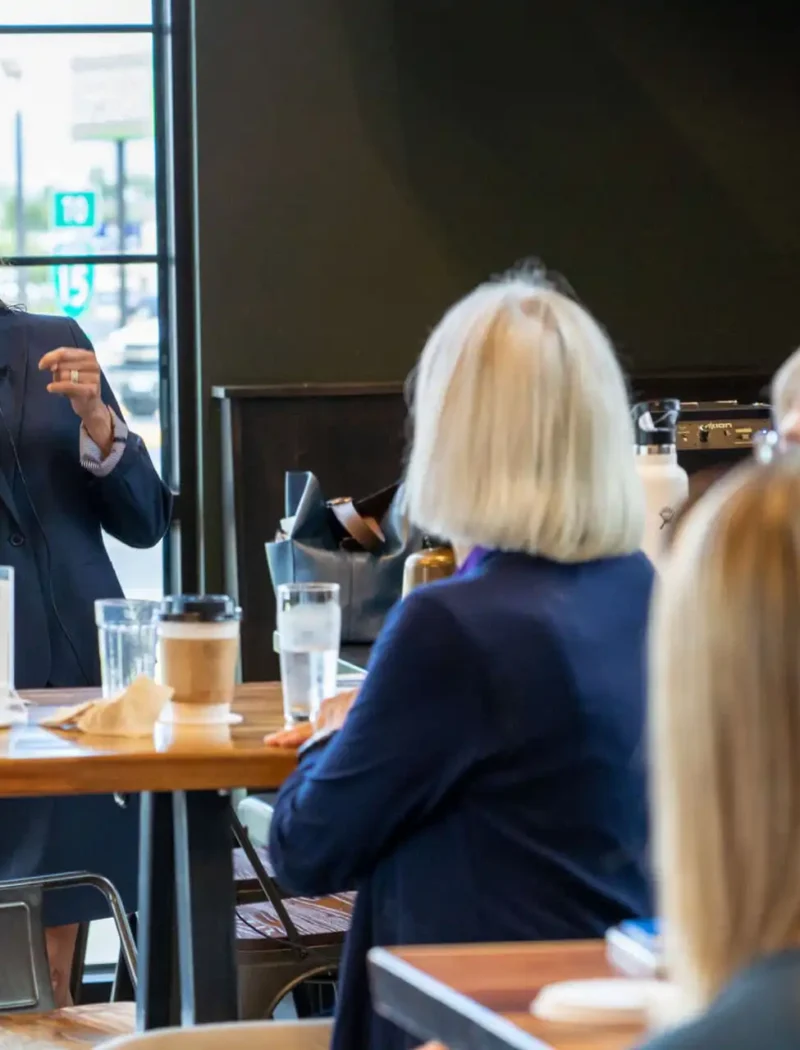 Secretary of State Christi Jacobsen smiles as she speaks into a microphone, addressing the Helena Rotary and a group of seated women in a bright, modern cafe with large windows.