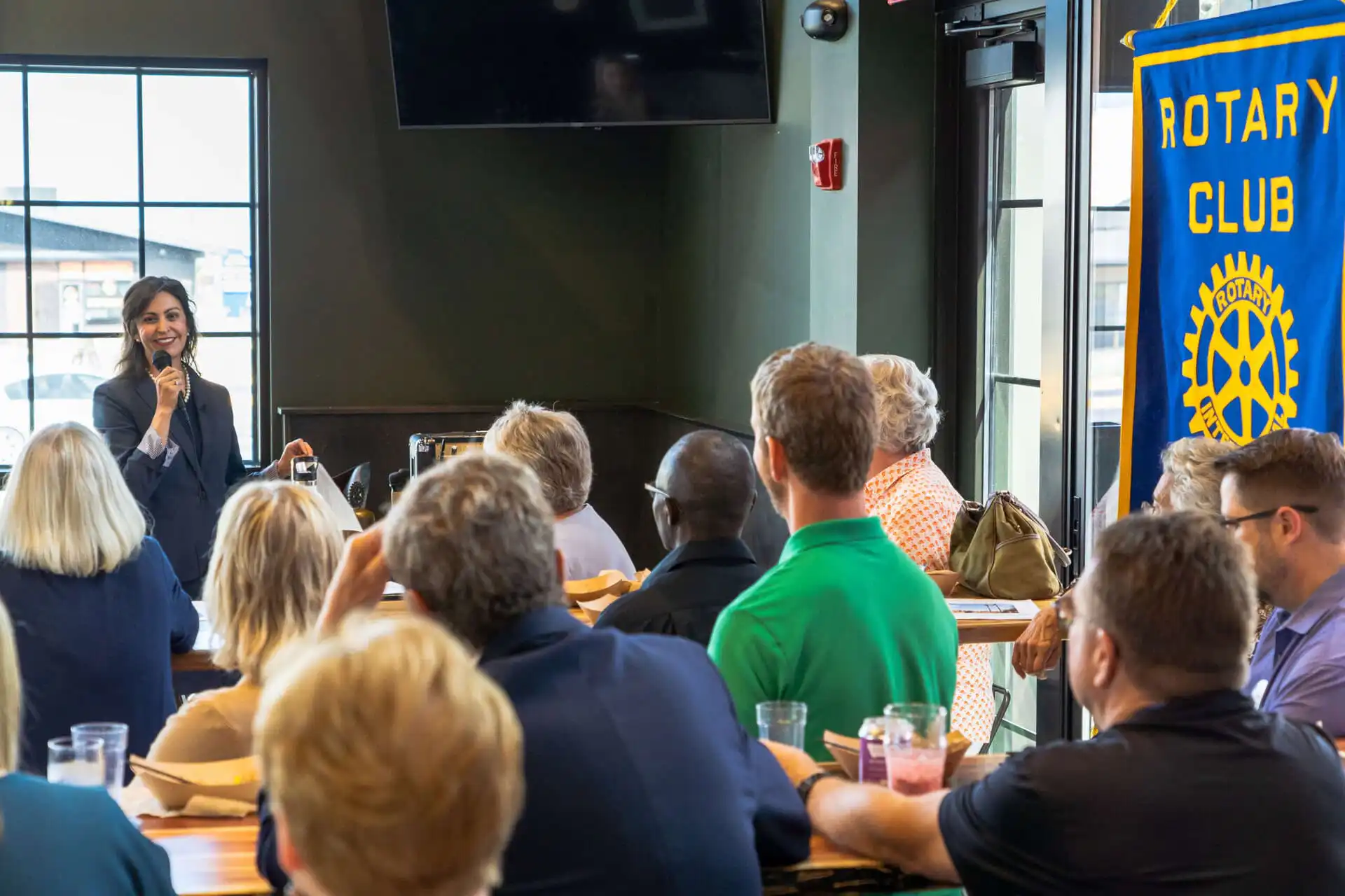 Christi Jacobsen, Montana Secretary of State, speaks to a group of people seated at tables during a Helena Rotary Club meeting in a room with large windows and a Rotary Club banner displayed.