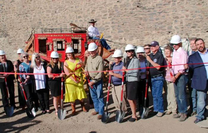Secretary Christi Jacobsen attends Stonewall Hall Phase 1 kickoff celebration event 3 A group of people in hard hats stand before Stonewall Hall with shovels, cutting a red ribbon at the kickoff celebration; Secretary Christi Jacobsen and a man on horseback are seen near the stagecoach and stone wall.