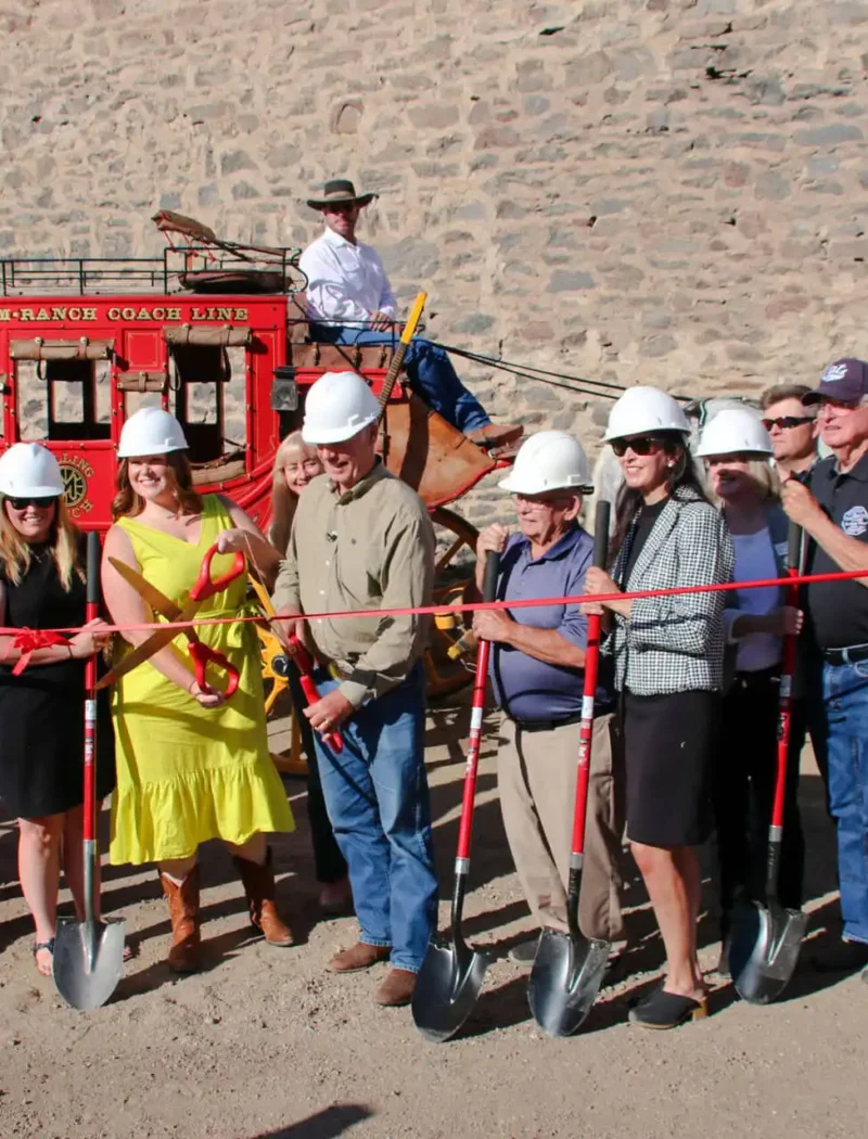Secretary Christi Jacobsen attends Stonewall Hall Phase 1 kickoff celebration event 6 A group of people in hard hats stand before Stonewall Hall with shovels, cutting a red ribbon at the kickoff celebration; Secretary Christi Jacobsen and a man on horseback are seen near the stagecoach and stone wall.