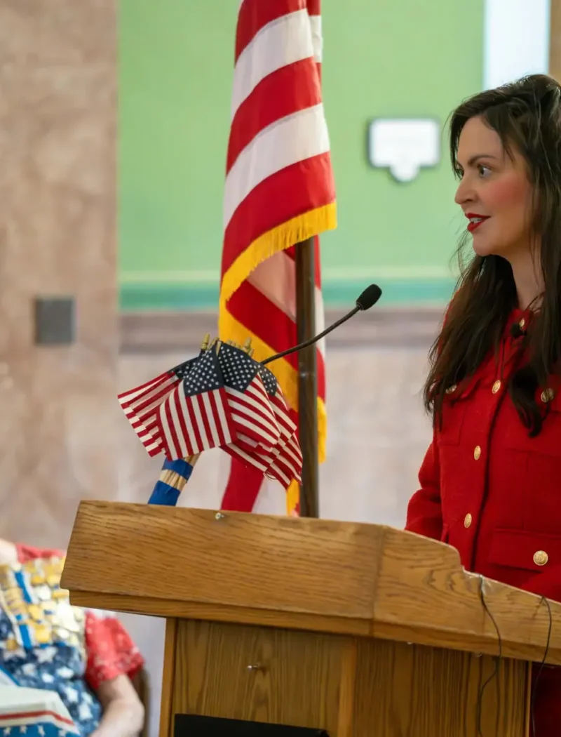 Christi Jacobsen, in a red coat, speaks at a podium decorated with small American flags during a Constitution Day celebration. An elderly woman sits beside her, while another woman in blue stands near an American flag in the background.