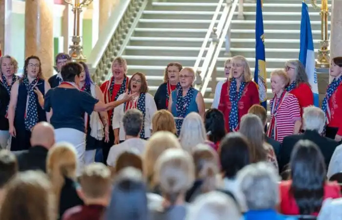 Secretary Christi Jacobsen joins Constitution Day celebration at Montana State Capitol 7 A womens choir dressed in red, white, and blue sings in front of an audience at the Montana State Capitol for Constitution Day, led by a conductor with flags and a grand staircase in the background. Secretary Christi Jacobsen attends the event.