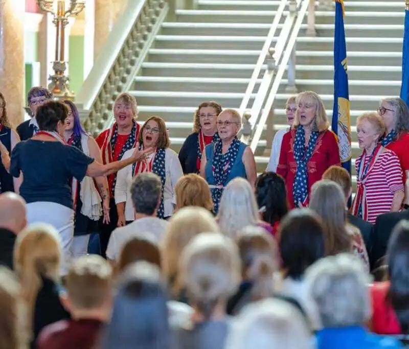 A womens choir dressed in red, white, and blue sings in front of an audience at the Montana State Capitol for Constitution Day, led by a conductor with flags and a grand staircase in the background. Secretary Christi Jacobsen attends the event.