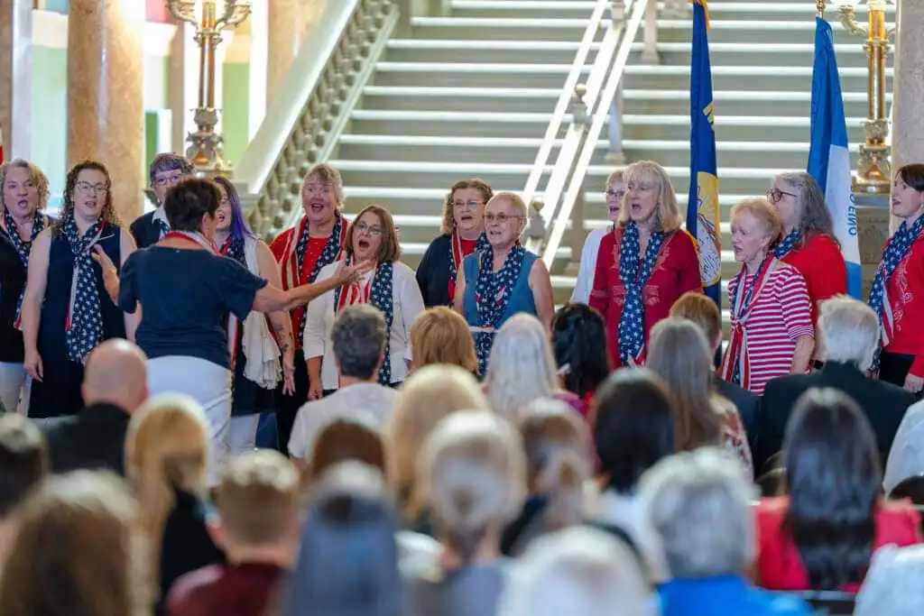 A womens choir dressed in red, white, and blue sings in front of an audience at the Montana State Capitol for Constitution Day, led by a conductor with flags and a grand staircase in the background. Secretary Christi Jacobsen attends the event.