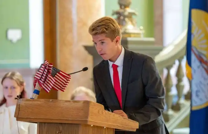 Secretary Christi Jacobsen announces 2023 Constitution Contest for Montana students 6 A young man in a suit and red tie speaks at a wooden podium with small American flags, participating in the Constitution Contest for Montana students, indoors with a flag and people in the background.