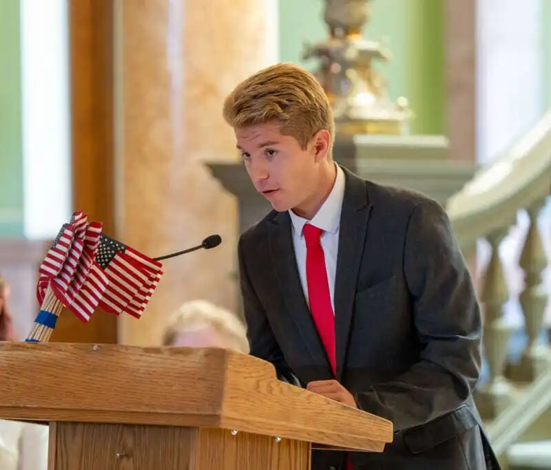 A young man in a suit and red tie speaks at a wooden podium with small American flags, participating in the Constitution Contest for Montana students, indoors with a flag and people in the background.