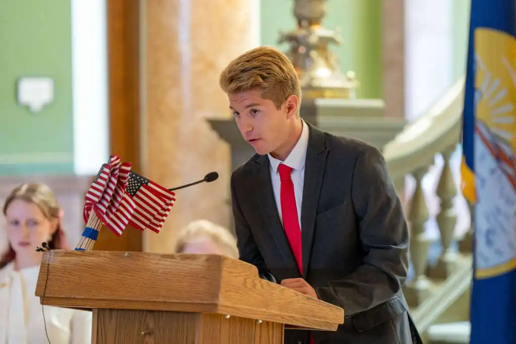 Secretary Christi Jacobsen announces 2023 Constitution Contest for Montana students 2 A young man in a suit and red tie speaks at a wooden podium with small American flags, participating in the Constitution Contest for Montana students, indoors with a flag and people in the background.