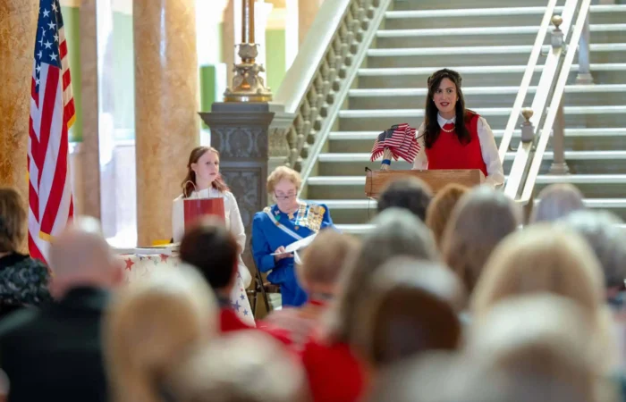 Secretary Christi Jacobsen announces 2023 Constitution Contest for Montana students 8 A woman speaks at a podium holding an American flag, with two women seated beside her, in front of a staircase and an audience. An American flag stands to the side.