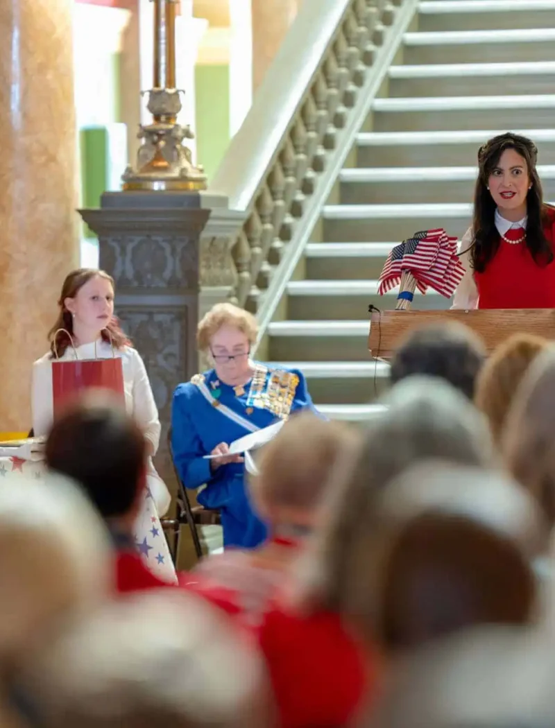 Secretary Christi Jacobsen announces 2023 Constitution Contest for Montana students 1 A woman speaks at a podium holding an American flag, with two women seated beside her, in front of a staircase and an audience. An American flag stands to the side.