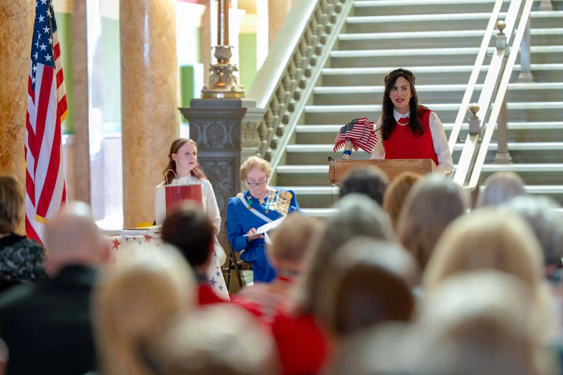 A woman speaks at a podium holding an American flag, with two women seated beside her, in front of a staircase and an audience. An American flag stands to the side.