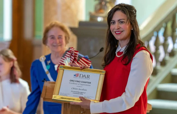 Secretary Christi Jacobsen joins Constitution Day celebration at Montana State Capitol 8 A smiling woman in a red dress holds a framed certificate, standing beside an older woman in a blue dress with a sash. Both are at a podium indoors, with another person blurred in the background.